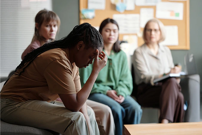 Young woman looking down, visibly distressed, with other individuals in the background, representing the experience of depression and the importance of depression treatment.