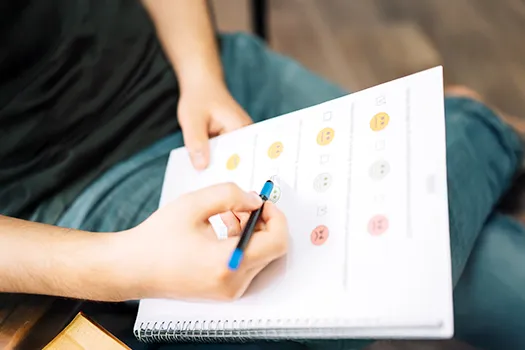 A person's hands filling out a mental health tracking sheet with emoji-like faces for mood ratings, signifying efficient mental health scheduling and patient progress monitoring.