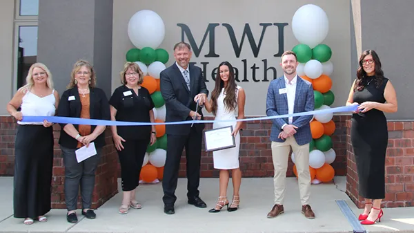 Grand reopening of MWI Health in Sioux Falls with a ribbon-cutting ceremony. The photo shows seven individuals, including MWI Health staff and local community leaders, smiling and holding a blue ribbon in front of the new clinic building. MWI Health's logo is visible behind them, along with green, orange, and white balloons.