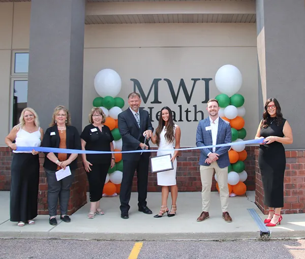 Ribbon-cutting ceremony for the new MWI Health facility in Rapid City, South Dakota. A group of seven individuals, including staff and community leaders, are shown cutting a blue ribbon in front of the clinic's MWI Health signage, flanked by green, orange, and white balloons.